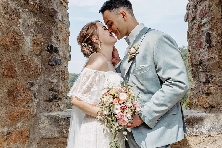 Braut und Bräutigam stehen eng beieinander und lächeln sich in romantischer Umarmung unter einem rustikalen Steinbogen an. Die Braut hält einen Strauß rosa-weißer Blumen in der Hand und trägt ein schulterfreies Kleid, der Bräutigam einen hellgrauen Anzug.