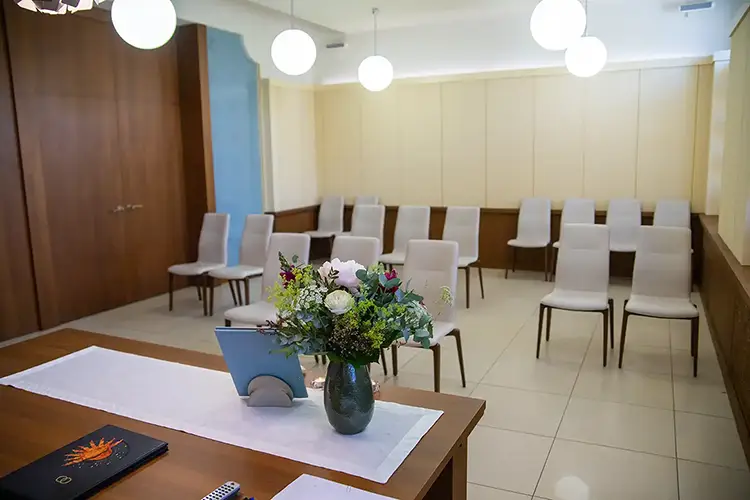 Ein kleiner Trauraum mit Reihen weißer Stühle, die an eine Hochzeit im Standesamt Radebeul erinnern, gegenüber einem mit Blumenarrangement und einem zugeklappten Laptop geschmückten Tisch. Kugelförmige Deckenleuchten beleuchten sanft die hölzernen und hellen Wände.