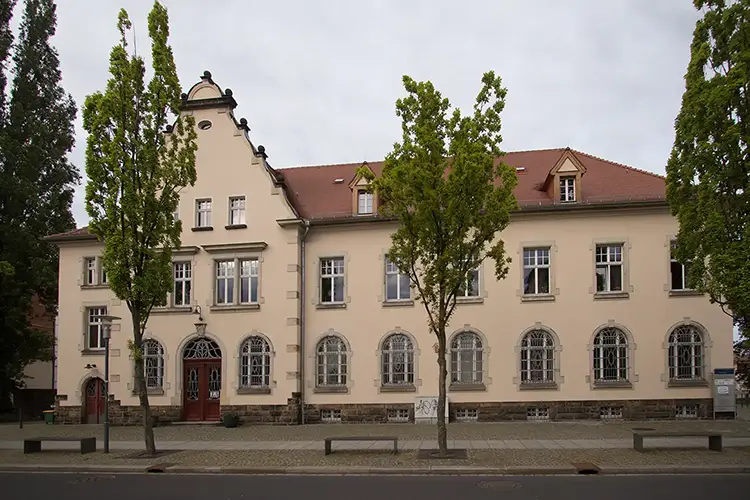 Hinter einem Bürgersteig steht ein historisches Gebäude mit beiger Fassade, rotem Dach und Bogenfenstern. Hohe, schmale Bäume säumen die Vorderseite, links befinden sich zwei rote Türen. Der Himmel ist bedeckt und bietet eine elegante Kulisse für alle, die im Standesamt Radebeul heiraten möchten.