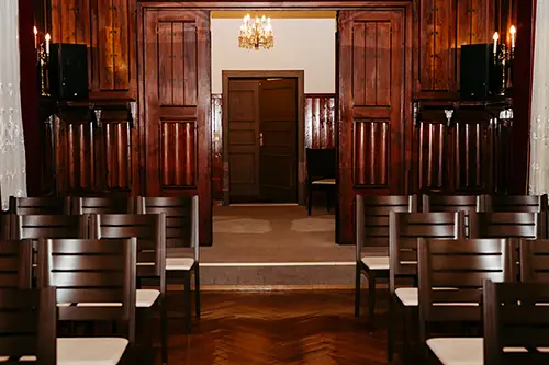 Ein holzgetäfelter Raum mit Reihen leerer Stühle vor einem erhöhten Podium bietet eine einladende Atmosphäre für die Hochzeit im Standesamt Tharandt. Doppeltüren öffnen sich zu einem schwach beleuchteten Flur, während ein Kronleuchter von Melanie Kunert Fotografie einen warmen Schein auf den polierten Boden wirft.