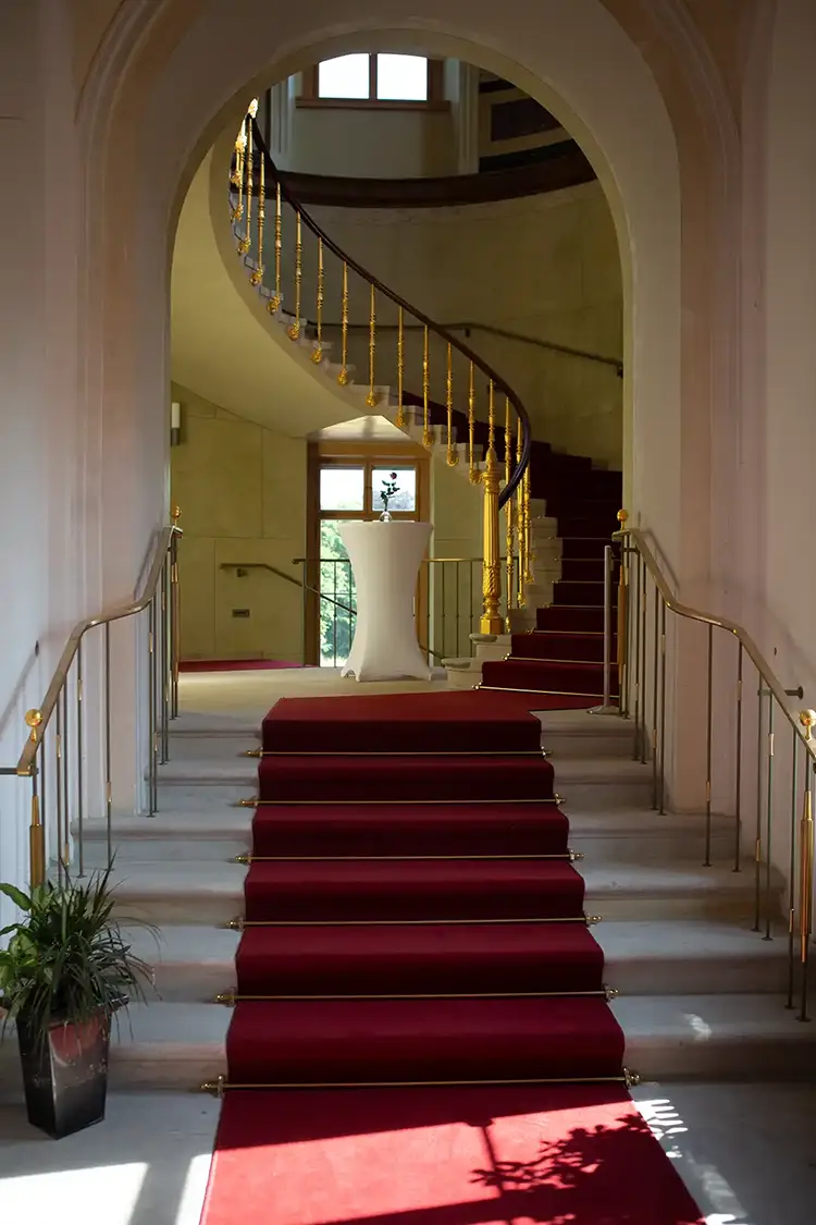 Eine große Treppe mit rotem Teppich und goldenem Geländer führt spiralförmig am Ende eines hellen Flurs hinauf – perfekt für eine Hochzeit im Lingnerschloss Dresden. Sonnenlicht fällt durch große Fenster und wirft Schatten an die Wände. Am Fuß der Treppe steht eine Topfpflanze.