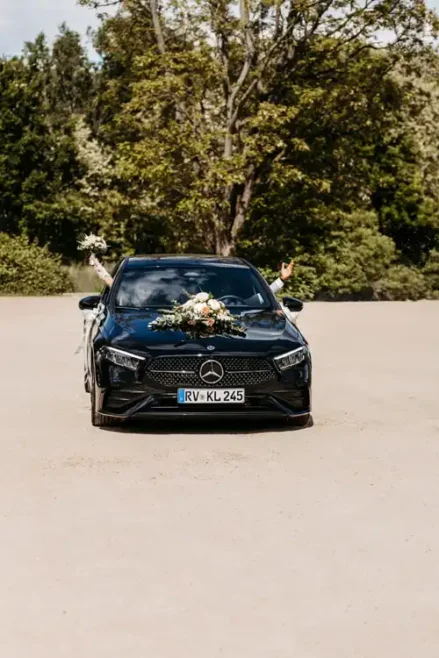 Ein schwarzer, mit Blumen geschmückter Mercedes parkt im Freien. Zwei Personen im Inneren strecken ihre Arme aus dem Fenster, jede hält einen Blumenstrauß - perfekt für eine Hochzeit auf Gut am See, wunderschön eingefangen von Hochzeitsfotograf Görlitz.