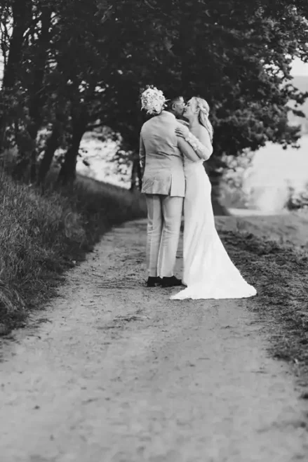 Ein Schwarz-Weiß-Foto eines Paares in Hochzeitskleidung, das sich auf einem von Bäumen gesäumten Weg auf Hochzeit Gut am See küsst, wobei die Braut ein langes Kleid trägt und der Bräutigam einen Blumenstrauß hinter dem Rücken hält. Wunderschön eingefangen von Hochzeitsfotograf Görlitz.