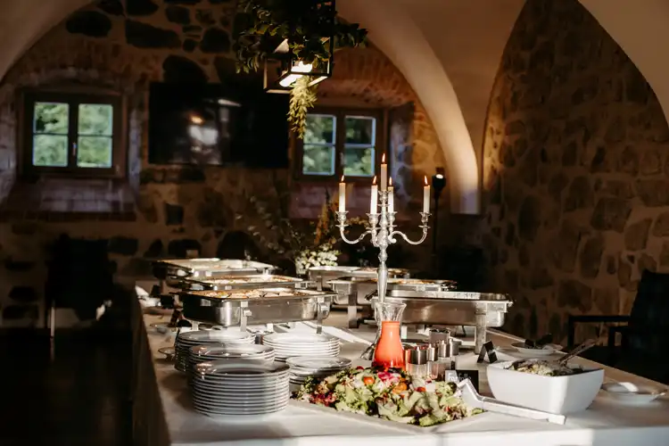 Ein Buffettisch in einem rustikalen Raum mit Steinmauern auf Hochzeit Gut am See, gedeckt mit Tellern, Besteck, Salaten, Chafing Dishes und einem beleuchteten silbernen Kandelaber unter gewölbten Decken und tageslichtdurchfluteten Fenstern.