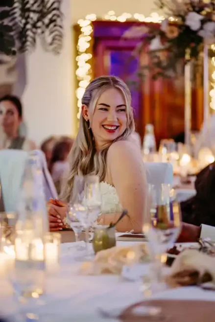 Eine Frau mit langen blonden Haaren in einem weißen Kleid lächelt strahlend, während sie an einem geschmückten Tisch während einer Hochzeit auf Gut am See sitzt, mit sanftem Kerzenlicht und Lichterketten im Hintergrund, aufgenommen von einem Hochzeitsfotograf Görlitz.