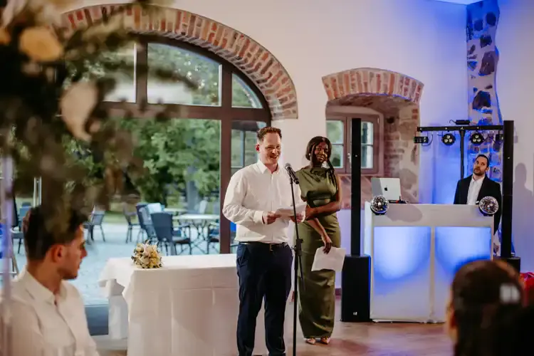 Auf einem Hochzeit Gut am See spricht ein Mann im weißen Hemd ins Mikrofon, während eine Frau in grünem Kleid lächelt. Im Hintergrund steht der DJ am beleuchteten Pult. Hochzeitsfotograf Görlitz hält die Gäste fest, die aufmerksam zuhören.
