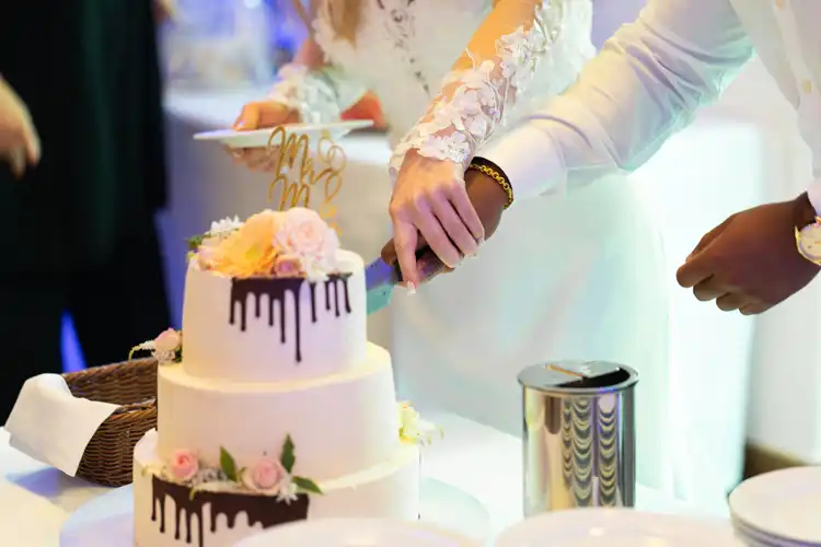 Braut und Bräutigam schneiden auf ihrer Hochzeit Gut am See eine weiße, zweistöckige Hochzeitstorte an, die mit Blumen und Schokoladentropfen verziert ist. Beide halten das Messer zusammen; die Braut trägt ein Kleid mit Spitzenärmeln. Auf dem Tisch stehen Teller und eine Dose.