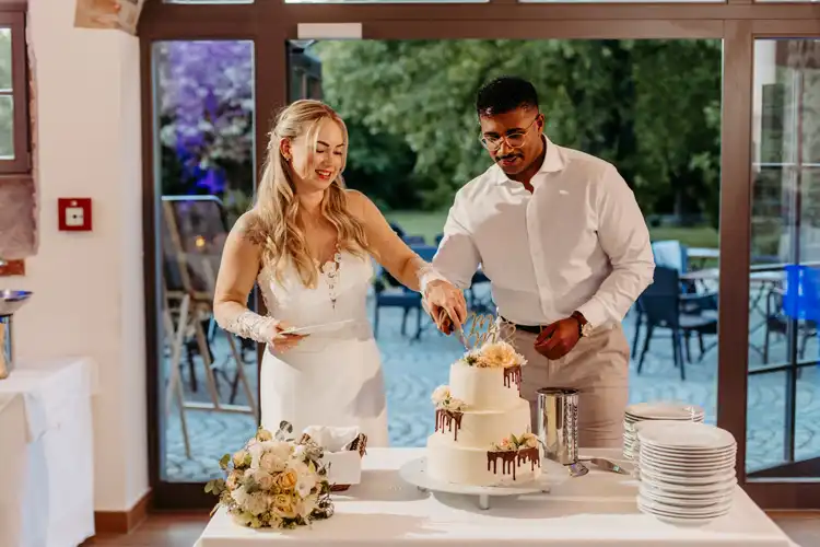 Eine Braut und ein Bräutigam stehen drinnen auf ihrer Hochzeit Gut am See zusammen und lächeln, während sie eine dreistöckige Hochzeitstorte anschneiden, die mit Blumen und Schokoladentropfen verziert ist. Auf dem Tisch stehen Teller und ein Blumenstrauß. Durch große Fenster kann man Grünpflanzen sehen.