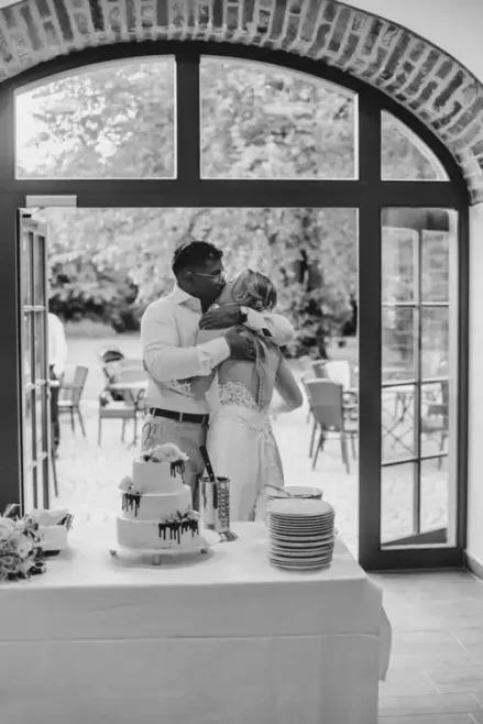 Ein Paar in Hochzeitskleidung umarmt sich neben einem Tisch mit einer dekorierten Torte und gestapelten Tellern, eingerahmt von gewölbten Backsteintüren auf Hochzeit Gut am See, mit Sitzgelegenheiten im Freien - wunderschön eingefangen von Hochzeitsfotograf Görlitz.