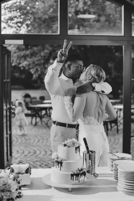 Ein frisch verheiratetes Paar küsst sich vor einem Fenster der Hochzeit Gut am See, wobei der Bräutigam ein Friedenszeichen macht. Eine dekorierte Hochzeitstorte und Teller stehen auf dem Tisch im Vordergrund. Das Bild ist in schwarz-weiß gehalten.