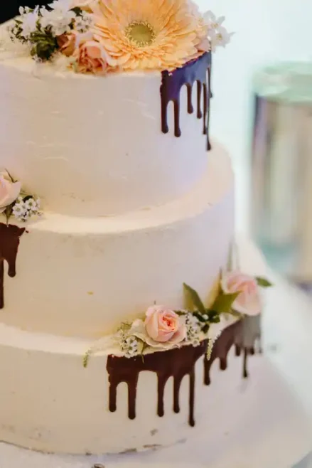 Eine zweistöckige weiße Torte, dekoriert mit Schokoladentropfen, kleinen rosafarbenen Rosen, weißen Blumen und Grünzeug, gekrönt von einer großen pfirsichfarbenen Blume - perfekt für eine Hochzeit auf Gut am See oder festgehalten von einem Hochzeitsfotografen Görlitz.