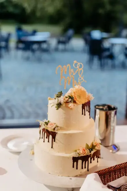 Eine dreistöckige weiße Hochzeitstorte mit Schokoladentropfen, die mit rosa und cremefarbenen Blumen und einem goldenen Mr & Mrs Topper verziert ist, ziert einen Tisch bei Hochzeit Gut am See; im Hintergrund sind verschwommene Tische und Stühle zu sehen.