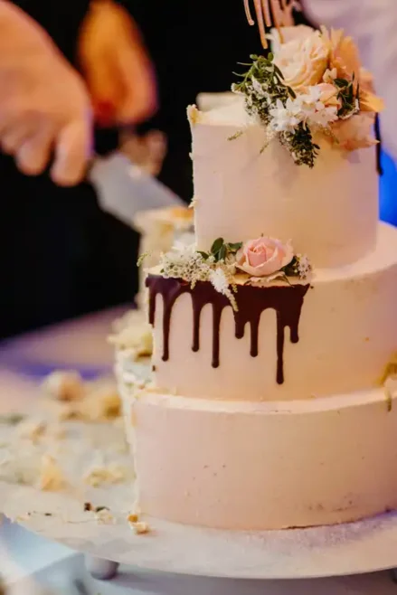 Auf einer Hochzeit Gut am See wird eine dreistöckige weiße Torte mit Schokodrip und rosa Blumen von einer Hand angeschnitten. Kuchenkrümel und Stücke liegen verstreut auf dem Tisch - festgehalten vom Hochzeitsfotograf Görlitz.