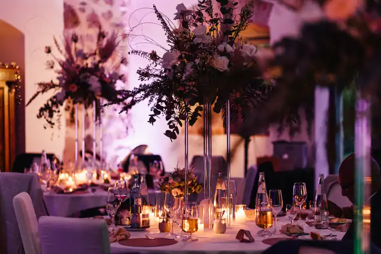 Eleganter Hochzeitsempfang auf Hochzeit Gut am See, mit runden Tischen, die mit hohen Blumenkränzen, leuchtenden Kerzen, Gläsern und sprudelndem Wasser dekoriert sind. Die romantische Atmosphäre wurde von Hochzeitsfotograf Görlitz wunderschön eingefangen.