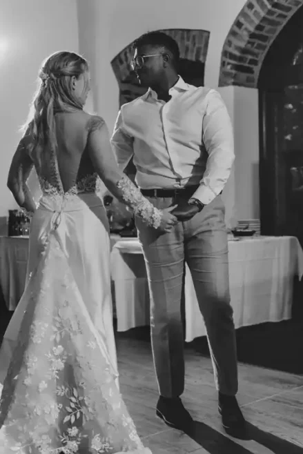 Eine Braut in einem rückenfreien geblümten Hochzeitskleid und ein Bräutigam in Hemd und Hose stehen eng beieinander auf der Tanzfläche der Hochzeit Gut am See, halten sich an den Händen und lächeln sich an. Eingefangen in zeitlosem Schwarz-Weiß von Hochzeitsfotograf Görlitz.