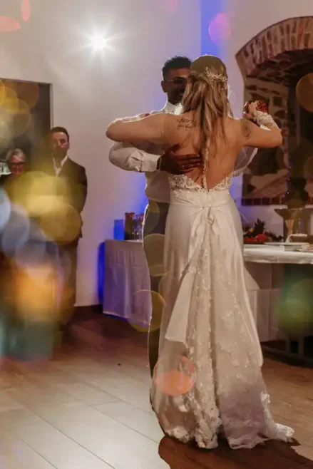 Eine Braut in einem weißen Kleid und ein Bräutigam tanzen ihren ersten Tanz auf ihrem Hochzeitsempfang auf Gut am See, während die Gäste zusehen und bunte Bokeh-Lichter im Vordergrund zu sehen sind.