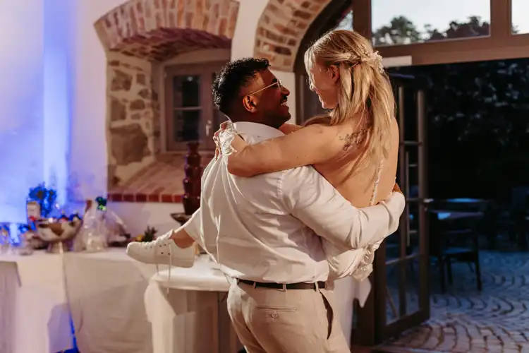 Ein Mann in einem weißen Hemd hebt eine Frau in einem weißen Kleid und Turnschuhen hoch und hält sie. Beide lächeln sich an in einem rustikalen, warm beleuchteten Raum mit Steinwänden und einem Desserttisch auf ihrer Hochzeit auf Gut am See.