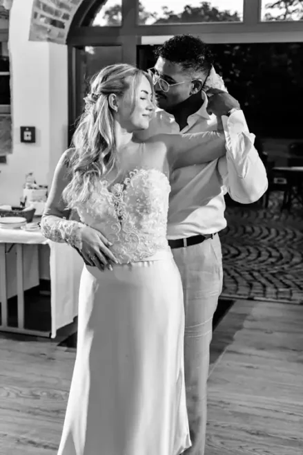 Ein Schwarz-Weiß-Foto eines Paares, das auf seiner Hochzeit auf Gut am See eng beieinander tanzt. Die Frau im Hochzeitskleid lächelt sanft und der Mann in Abendgarderobe steht hinter ihr und hält sanft ihren Arm.