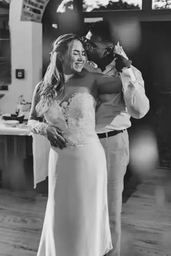 Eine lächelnde Braut im Spitzenhochzeitskleid wird von ihrem Partner von hinten umarmt, der sie auf die Schläfe küsst. Das glückliche Paar, fotografiert von Hochzeitsfotograf Görlitz, teilt einen entspannten Moment in einer warm beleuchteten Innenumgebung. Das Foto ist in schwarz-weiß gehalten.