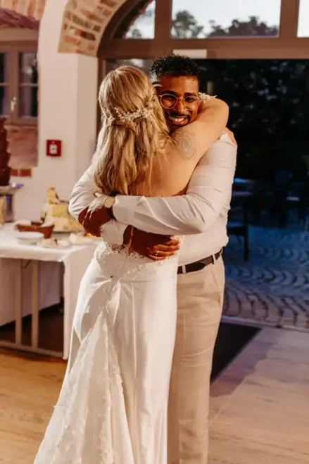 Eine Braut in einem weißen Kleid umarmt einen lächelnden Mann in einem weißen Hemd und einer beigen Hose in einem Innenraum von Hochzeit Gut am See, mit einem Banketttisch und einem gewölbten Fenster im Hintergrund.