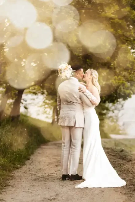 Eine Braut und ein Bräutigam stehen auf einem von Bäumen gesäumten Weg bei Hochzeit Gut am See, umarmen sich und lächeln sich an. Die Braut hält einen Blumenstrauß, der Bräutigam trägt einen leichten Anzug. Sanfte goldene Bokeh-Lichter erzeugen einen verträumten Effekt.