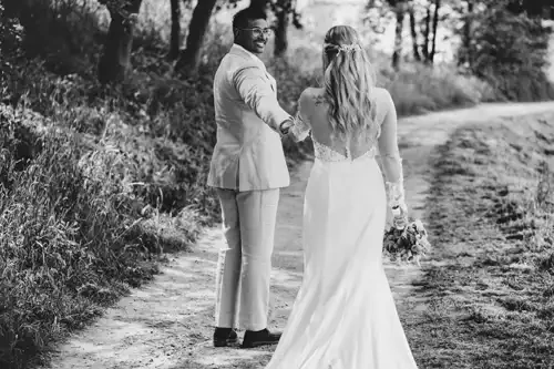 Eine Braut und ein Bräutigam gehen Hand in Hand einen Waldweg bei Hochzeit Gut am See hinunter. Der Bräutigam lächelt zurück in die Kamera, während die Braut mit ihrem Brautstrauß in der Hand das Gesicht abwendet. Dieser Schwarz-Weiß-Moment wurde vom Hochzeitsfotograf Görlitz wunderschön eingefangen.