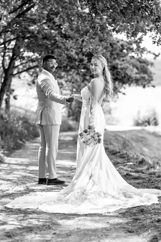 Eine Braut in einem langen, eleganten Kleid hält einen Blumenstrauß und blickt lächelnd zurück, während sie die Hand des Bräutigams hält. Sie stehen zusammen auf einem sonnenbeschienenen, von Bäumen gesäumten Weg - wunderschön eingefangen vom Hochzeitsfotograf Görlitz. Das Foto ist in schwarz-weiß gehalten.