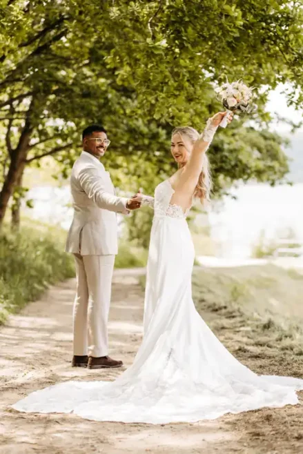 Eine Braut in einem weißen Kleid hält einen Blumenstrauß in der Hand und hebt freudig einen Arm, um in die Kamera zu lächeln. Ihr Bräutigam steht neben ihr, Hand in Hand, auf einem sonnenbeschienenen Weg - eingefangen von Hochzeitsfotograf Görlitz auf Hochzeit Gut am See.