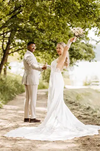 Eine Braut in einem weißen Kleid hält einen Blumenstrauß in der Hand und hebt freudig einen Arm, um in die Kamera zu lächeln. Ihr Bräutigam steht neben ihr, Hand in Hand, auf einem sonnenbeschienenen Weg - eingefangen von Hochzeitsfotograf Görlitz auf Hochzeit Gut am See.