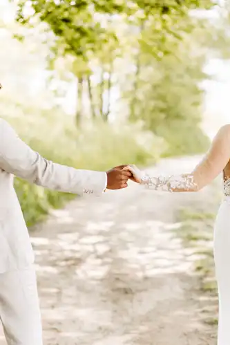 Ein händchenhaltendes Paar im Freien auf einem sonnenbeschienenen Feldweg, in heller Festtagskleidung; nur die Arme und ein Teil des Körpers sind zu sehen - aufgenommen von Hochzeitsfotograf Görlitz auf Hochzeit Gut am See.