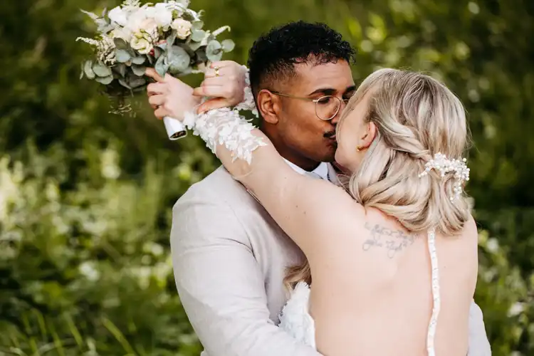 Eine Braut und ein Bräutigam küssen sich im Freien auf der Hochzeit Gut am See. Die Braut hebt ihren Brautstrauß und enthüllt ein Tattoo auf ihrer Schulter, während der Bräutigam mit Brille und hellem Anzug inmitten von üppigem Grün steht. Aufgenommen von Hochzeitsfotograf Görlitz.