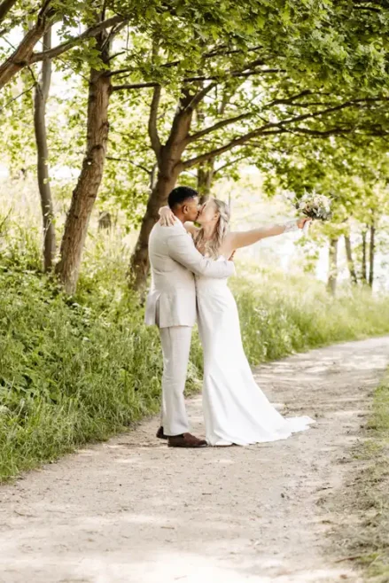 Braut und Bräutigam küssen sich auf einem von Bäumen gesäumten Weg bei Hochzeit Gut am See. Die Braut hält einen Blumenstrauß in die Höhe, während das Sonnenlicht durch die grünen Blätter fällt und eine romantische, natürliche Umgebung schafft, die von Hochzeitsfotograf Görlitz eingefangen wurde.