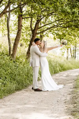 Braut und Bräutigam küssen sich auf einem von Bäumen gesäumten Weg bei Hochzeit Gut am See. Die Braut hält einen Blumenstrauß in die Höhe, während das Sonnenlicht durch die grünen Blätter fällt und eine romantische, natürliche Umgebung schafft, die von Hochzeitsfotograf Görlitz eingefangen wurde.
