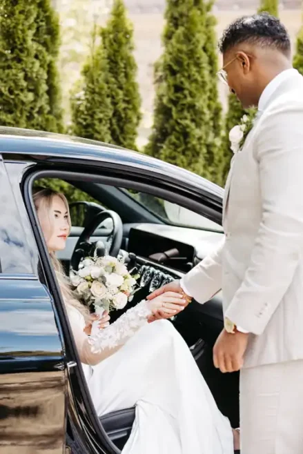 Eine Braut in einem weißen Kleid sitzt in einem Auto und hält einen Strauß weißer Blumen, während ihr Bräutigam draußen sanft ihre Hand hält. Grüne Bäume und die romantische Kulisse von Hochzeit Gut am See sorgen für perfekte Erinnerungen von Hochzeitsfotograf Görlitz.