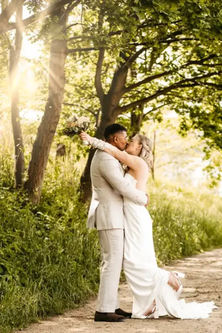 Eine Braut und ein Bräutigam küssen sich auf einem sonnenbeschienenen, von Bäumen gesäumten Weg bei Hochzeit Gut am See. Die Braut hebt ihren Brautstrauß, das Sonnenlicht bricht durch die Blätter, während ein Hochzeitsfotograf Görlitz diesen magischen Moment festhält.