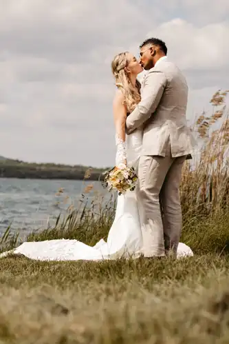 Braut und Bräutigam küssen sich am See bei der Hochzeit Gut am See. Die Braut hält einen Blumenstrauß in ihrem weißen Kleid, während der Bräutigam einen hellen Anzug trägt. Das hohe Gras und das glitzernde Wasser bilden eine romantische Kulisse.
