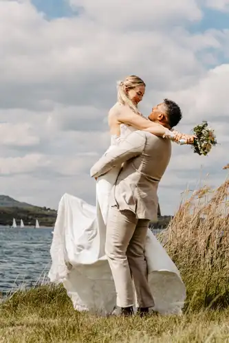 Ein Bräutigam hebt seine Braut am Seeufer von Hochzeit Gut am See in die Arme, beide lächeln freudig. Die Braut hält einen Blumenstrauß in der Hand und trägt Weiß, während Segelboote und Hügel den Hintergrund unter einem teilweise bewölkten Himmel zieren.