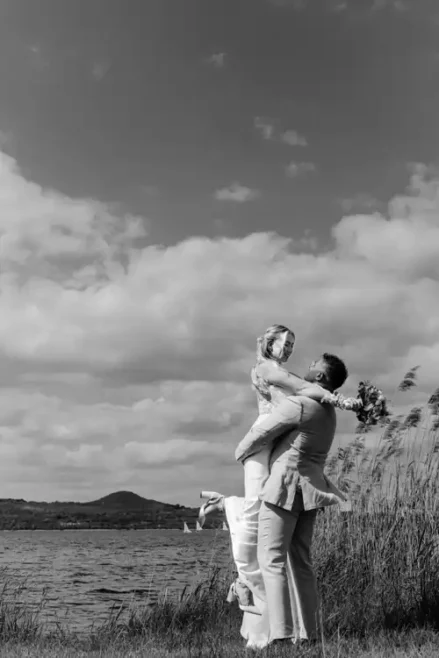 Ein Bräutigam hebt seine Braut am Seeufer von Hochzeit Gut am See in die Arme, beide lächeln freudig. Im Freien, mit Schilf, Wasser und Hügeln im Hintergrund unter einem teilweise bewölkten Himmel. Das Schwarz-Weiß-Foto fängt das pure Glück ein.
