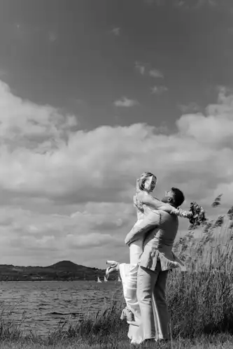 Ein Bräutigam hebt seine Braut am Seeufer von Hochzeit Gut am See in die Arme, beide lächeln freudig. Im Freien, mit Schilf, Wasser und Hügeln im Hintergrund unter einem teilweise bewölkten Himmel. Das Schwarz-Weiß-Foto fängt das pure Glück ein.