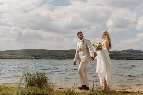 Eine Braut und ein Bräutigam gehen Hand in Hand am Seeufer von Hochzeit Gut am See spazieren und lächeln sich an. Der Bräutigam trägt einen hellen Anzug und die Braut hält einen Blumenstrauß. Im Hintergrund sind grüne Hügel und ein bewölkter Himmel zu sehen.