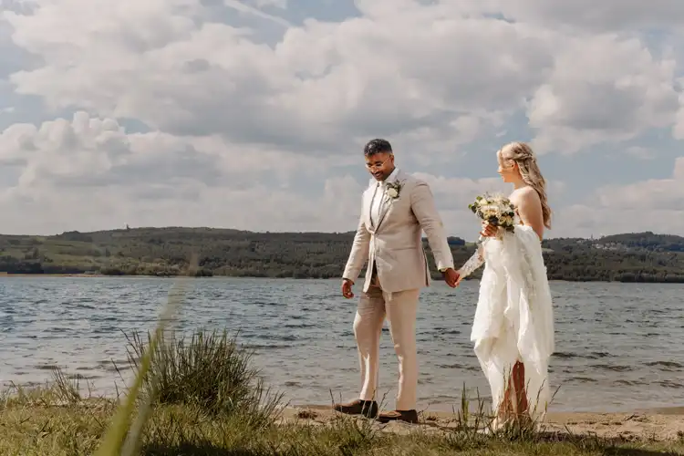 Braut und Bräutigam spazieren Hand in Hand am Seeufer von Hochzeit Gut am See entlang, gekleidet in Hochzeitskleidung, mit grasbewachsenen Ufern und Hügeln im Hintergrund unter einem teilweise bewölkten Himmel.