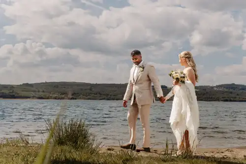 Braut und Bräutigam spazieren Hand in Hand am Seeufer von Hochzeit Gut am See entlang, gekleidet in Hochzeitskleidung, mit grasbewachsenen Ufern und Hügeln im Hintergrund unter einem teilweise bewölkten Himmel.