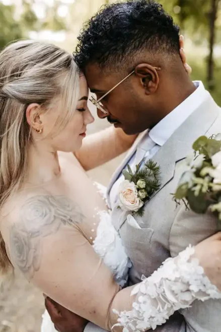 Eine Braut und ein Bräutigam stehen eng beieinander im Freien auf der Hochzeit Gut am See, berühren sich an der Stirn und umarmen sich. Die Braut hält einen Blumenstrauß in der Hand und hat ein Blumentattoo auf dem Arm; beide wirken glücklich und friedlich. Aufgenommen von Hochzeitsfotograf Görlitz.