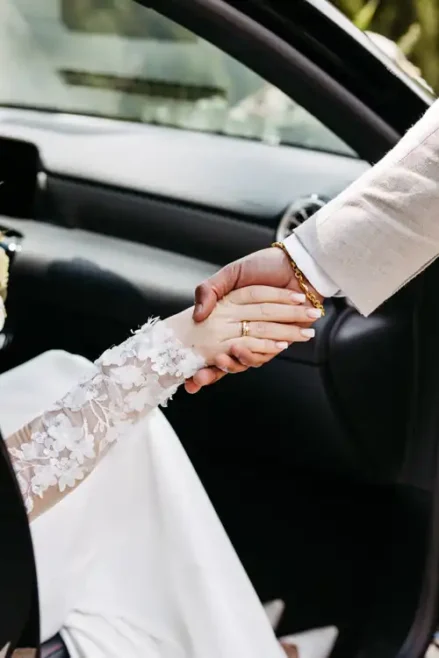 Eine Braut in einem weißen, geblümten Spitzenärmel hält die Hand ihres Partners in einem beigen Anzug, während sie aus dem Auto aussteigt - ein intimer Moment, der von Hochzeitsfotograf Görlitz auf Hochzeit Gut am See festgehalten wurde.
