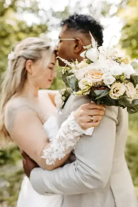 Braut und Bräutigam umarmen sich im Freien auf Hochzeit Gut am See. Der Bräutigam küsst die Stirn der Braut, die einen Strauß weißer und pfirsichfarbener Blumen in der Hand hält, umgeben von Grünpflanzen, durch die das Sonnenlicht scheint. Hochzeitsfotograf Görlitz hält den Moment fest.