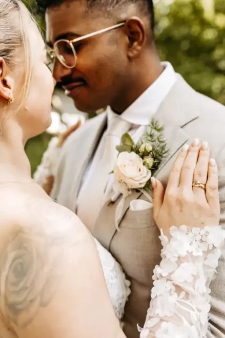 Braut und Bräutigam stehen bei ihrer Hochzeit auf Gut am See eng beieinander und blicken sich in die Augen. Die Hand der Braut mit Ehering ruht auf seiner Schulter, der Bräutigam, im hellen Anzug und mit rosa Ansteckblume, trägt eine Brille. Hochzeitsfotograf Görlitz hält den Moment fest.