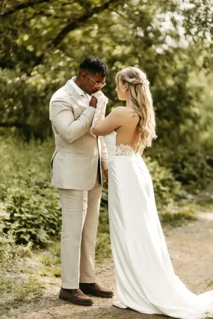 Ein Bräutigam in einem beigen Anzug küsst sanft die Hand seiner Braut in Spitze, während sie auf einem Weg im Grünen stehen. Dieser intime Moment im Freien wurde von Hochzeitsfotograf Görlitz auf Hochzeit Gut am See wunderschön eingefangen.