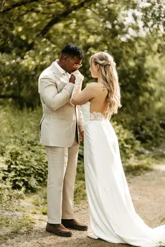 Ein Bräutigam in einem beigen Anzug küsst sanft die Hand seiner Braut in Spitze, während sie auf einem Weg im Grünen stehen. Dieser intime Moment im Freien wurde von Hochzeitsfotograf Görlitz auf Hochzeit Gut am See wunderschön eingefangen.