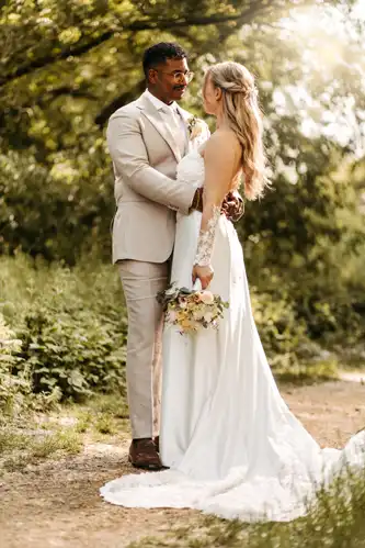 Eine Braut und ein Bräutigam stehen eng beieinander im Freien auf Hochzeit Gut am See und schauen sich in die Augen. Die Braut hält einen Blumenstrauß, der Bräutigam einen leichten Anzug, während das Sonnenlicht durch die Bäume fällt - wunderschön eingefangen von Hochzeitsfotograf Görlitz.