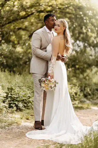 Eine Braut in einem weißen Kleid hält einen Blumenstrauß, während sie mit einem Bräutigam in einem hellen Anzug steht. Sie umarmen sich im Freien auf einem sonnenbeschienenen Weg auf Hochzeit Gut am See, umgeben von Grün, und wirken friedlich und glücklich. Hochzeitsfotograf Görlitz hat den Moment festgehalten.